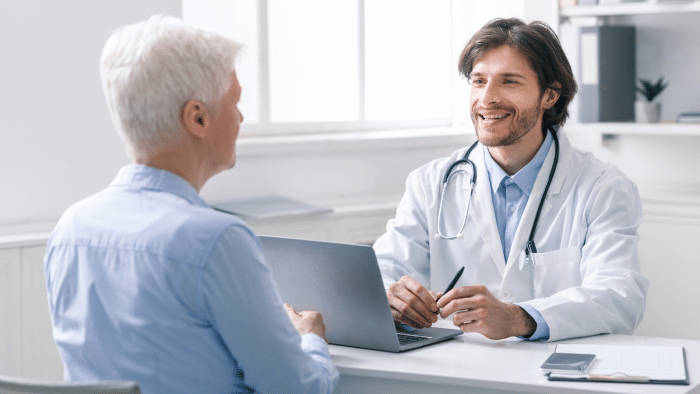 A doctor with a stethoscope around his neck sits at a desk in an office, smiling while talking to an elderly patient with white hair. The doctor is holding a pen and there is a laptop, a notepad, and a phone on the desk.