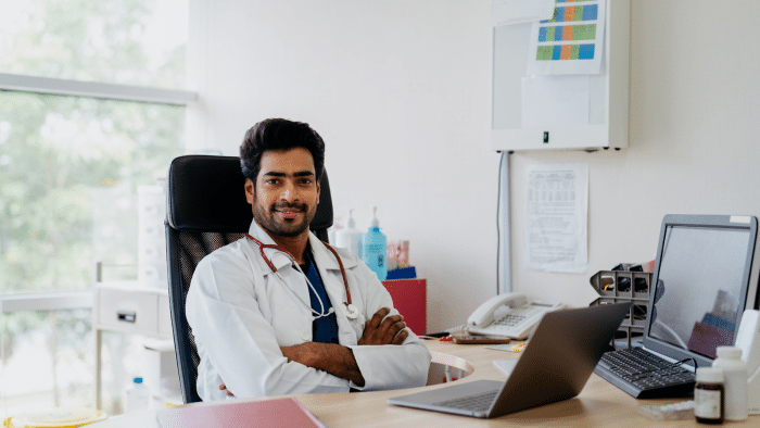 A doctor wearing a white coat and stethoscope sits confidently at a desk with a laptop, phone, and various office supplies. The background features a large window with a blurry outdoor view, paperwork on a bulletin board, and medical equipment nearby.