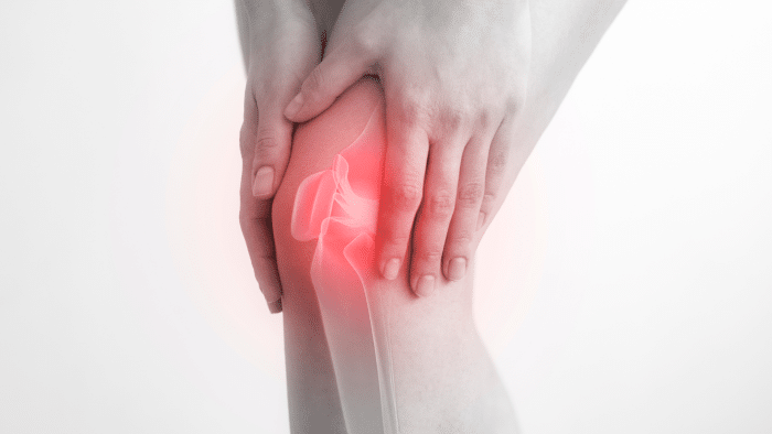 A person holds their knee with both hands, suggesting discomfort or injury. The knee area is highlighted with a red glow, indicating pain or inflammation. The image has a grayscale background with the red glow providing a stark contrast.