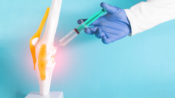 A gloved hand injects a syringe into a model of a human knee joint, highlighted with yellow ligaments. The area around the needle is glowing red, suggesting inflammation or pain treatment. The background is blue.