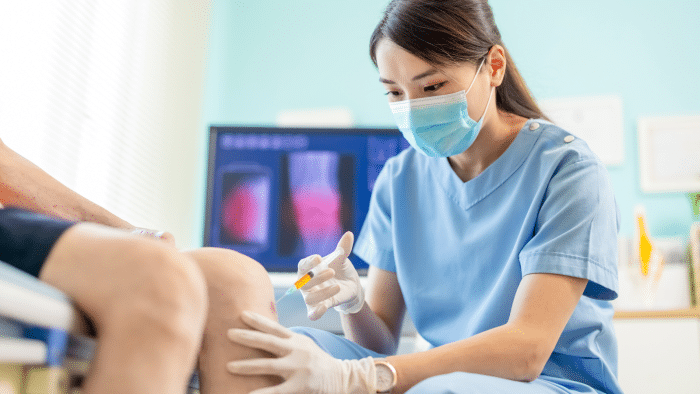 A healthcare professional wearing blue scrubs, a face mask, and gloves administers an injection to a patient's knee. In the background, an X-ray of a knee is displayed on a computer screen. The setting appears to be a bright, modern medical office.