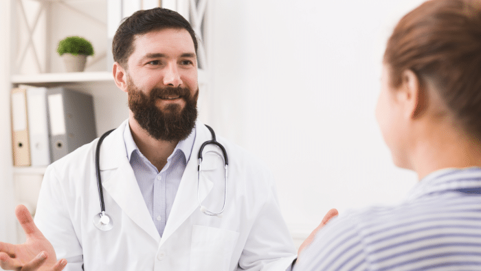 A bearded doctor in a white coat and stethoscope is smiling and talking to a patient. The patient is seen from behind. The scene is set in a medical office with shelves and binders in the background, conveying a professional and approachable atmosphere.