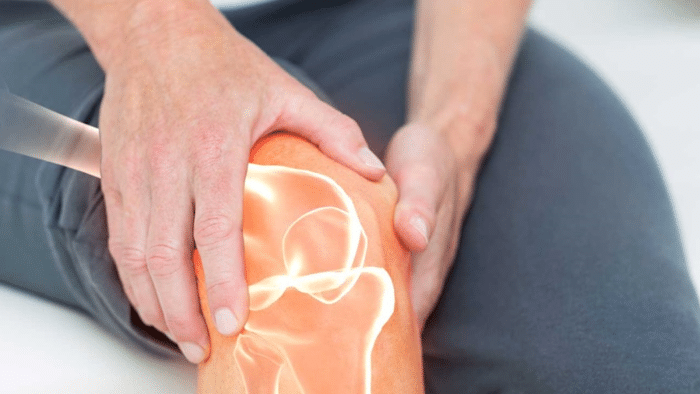 A person holding their knee with both hands, indicating pain or discomfort. An illustration of bones and joints is superimposed over the knee, suggesting the person is experiencing joint or knee pain. The background is slightly blurred.