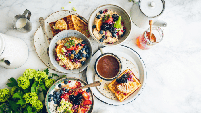 A breakfast spread on a white marble surface includes waffles topped with berries, bowls of oatmeal garnished with fruit, a cup of coffee, a jar of honey, a small pitcher of milk, and a white teapot. A bunch of green hydrangeas adorns the corner of the table.