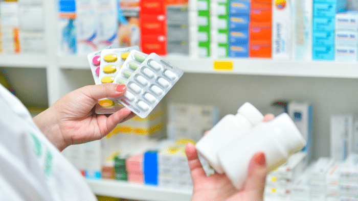 A person holds blister packs of pills and white pill bottles in a pharmacy. The background shows shelves filled with various medications and boxes. The person is wearing a white coat, likely indicating a pharmacist.