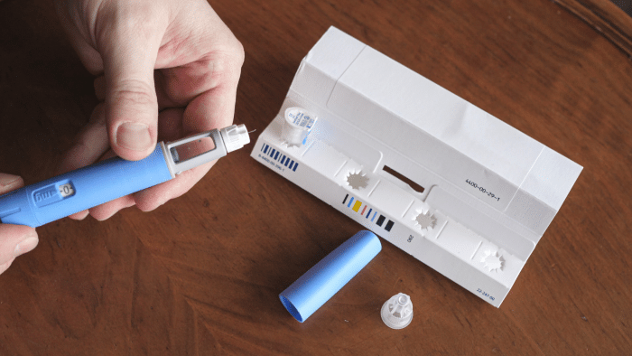 A person is assembling a blue insulin pen next to an opened packaging box on a wooden surface. The packaging includes various components of the insulin pen laid out, including the pen cap and needle tips. The person's hands are visible, holding the pen body.