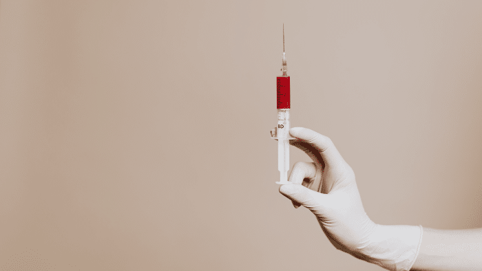 A gloved hand holding a syringe filled with red liquid against a neutral background. The syringe is oriented vertically with the needle pointing upwards.