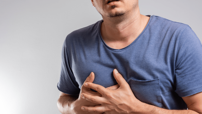 Young man in a blue T-shirt clutching his chest with both hands, suggesting he may be experiencing chest pain or discomfort. The background is plain and light gray. His face is partially visible but not the focal point.