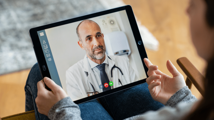 A person holds a tablet showing a video call with a doctor. The doctor, wearing a white coat and stethoscope, appears focused and engaged. The person holds the tablet in both hands, participating in a telehealth consultation.