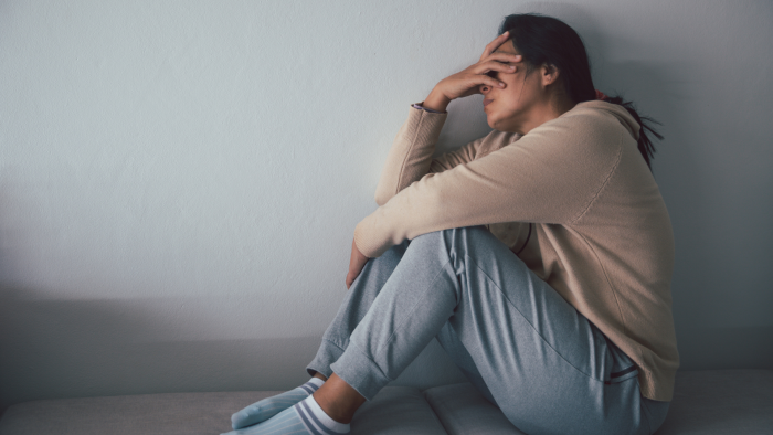 A person wearing a beige sweater and light gray sweatpants sits on a couch with their legs drawn up, resting their head on their hand in a gesture of frustration or exhaustion. The background is a plain, white wall.