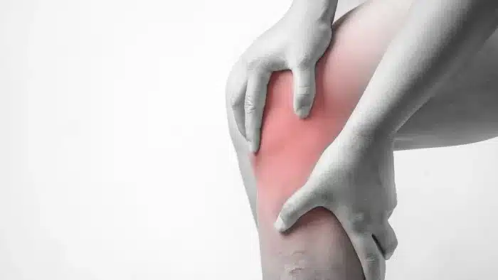 A grayscale image showing a person holding their knee, highlighted in red to indicate pain or injury. The hands are positioned on either side of the knee, suggesting discomfort or a need for support in that area.