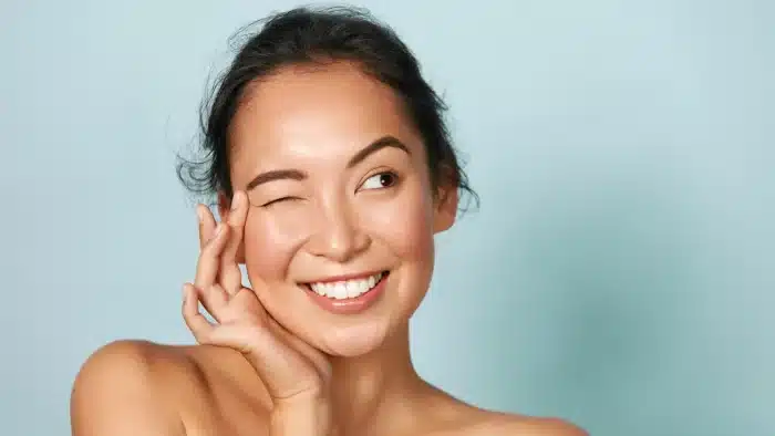 A smiling person winks with their right eye, softly touching their right cheek with their right hand. They have dark hair pulled back, and their skin looks smooth and radiant against a light blue background. The person appears joyful and relaxed.