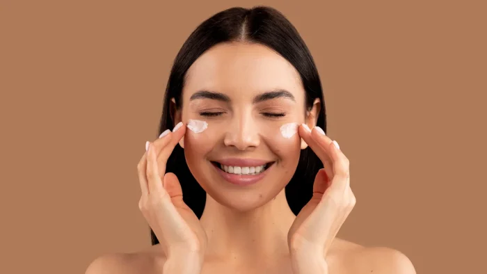 A woman with long brown hair and closed eyes is smiling while applying two dabs of cream to her cheeks. She is using her fingertips to spread the cream. The background is a solid light brown color.