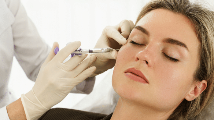 A woman with closed eyes receives a cosmetic injection from a medical professional wearing white gloves. The injection is administered to the area near her cheek. The setting appears to be a clinical or spa environment.