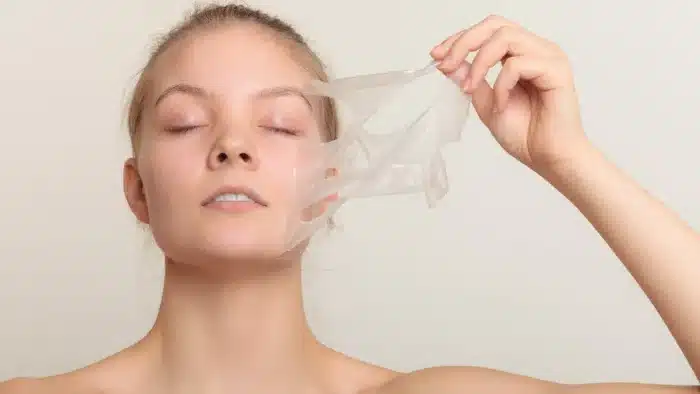 A woman with closed eyes gently peels off a transparent facial mask from the left side of her face with her right hand. She appears relaxed and serene, with a neutral beige background.