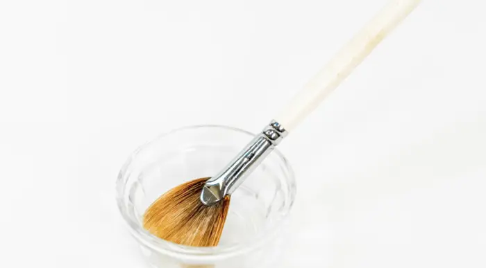 A wooden-handled paintbrush with brown bristles is resting inside a small, clear glass bowl. The brush is angled, and its bristles are slightly fanned out. The background is plain white.