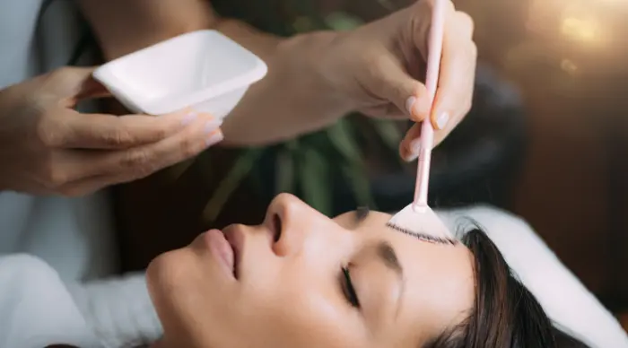 A person with closed eyes is receiving a facial treatment. A hand is applying a cosmetic mask or cream to their forehead with a brush, while the other hand holds a small white bowl. The scene suggests a relaxing skincare or spa environment.