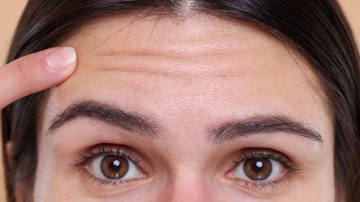 Close-up of a person's face focusing on their forehead, eyes, and eyebrows. Their right hand points to the forehead, highlighting fine lines and wrinkles. The person has well-groomed eyebrows and natural skin texture.