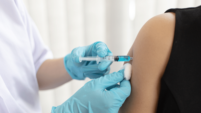 A healthcare professional wearing blue gloves is administering a vaccine into a person's arm using a syringe. The healthcare worker is holding a cotton ball near the injection site. The person receiving the vaccine is wearing a sleeveless black top.