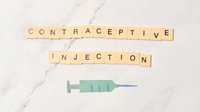 A marble surface with Scrabble tiles spelling "CONTRACEPTIVE INJECTION" arranged in two lines. Below the text is a flat paper cutout of a syringe in blue and green.