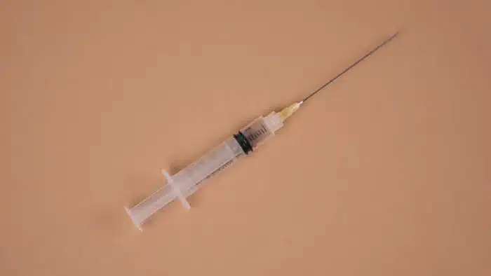 A clear plastic syringe with an attached needle is placed on a light brown surface. The syringe is empty, showing the plunger in a rest position and marked with measurement lines. The needle cap is off, exposing the sharp needle tip.