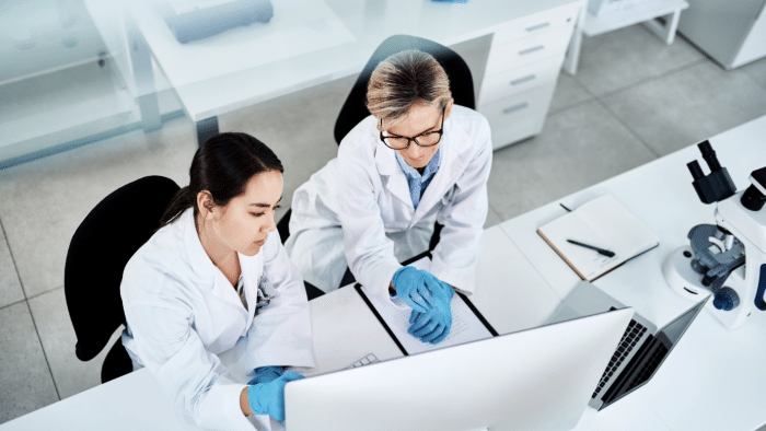 Two scientists in lab coats and gloves are working together at a desk with a computer monitor and a laptop in a bright laboratory. One scientist is pointing at the screen while the other takes notes. Lab equipment, including a microscope, is visible in the background.