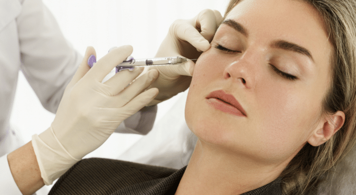 A woman with closed eyes is receiving an injection in her cheek from a medical professional wearing gloves and holding a syringe. The procedure appears to be a cosmetic treatment.