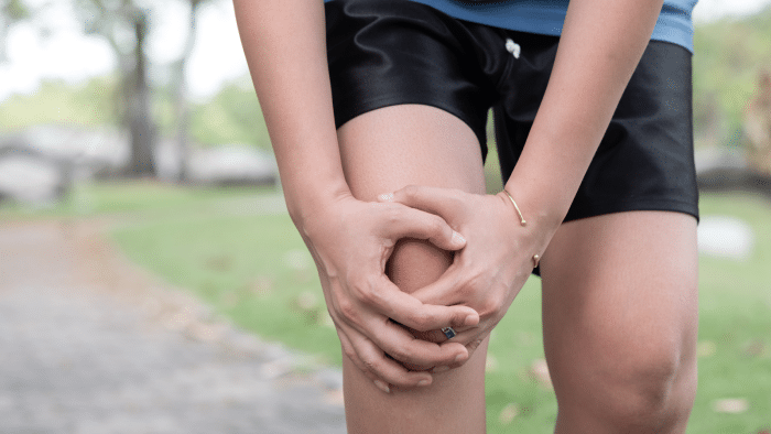 Person clutching their knee with both hands, indicating pain or discomfort. They are outdoors on a paved path with green grass and trees in the background, wearing black shorts and a blue top. The image suggests a possible knee injury or strain during physical activity.