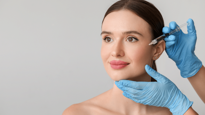 A calm woman with brown hair receives a cosmetic injection in her cheek from a professional wearing blue gloves. The professional steadies the woman's chin with one hand while administering the injection with the other. The background is a plain, light gray color.