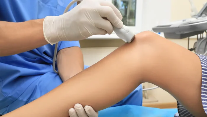 A healthcare professional in blue scrubs performs an ultrasound scan on a patient's knee. The professional is wearing white gloves and holding the ultrasound probe against the knee. The patient lies down with their leg extended.