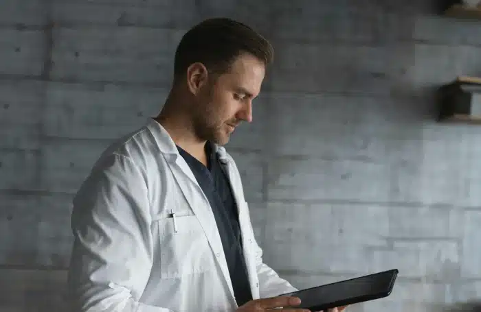 A male doctor wearing a white lab coat and dark clothing underneath is standing and looking at a tablet in his hands. He is in a modern, minimalistic room with a concrete-textured wall in the background. The atmosphere is professional and focused.