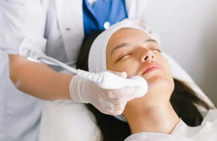 A person is lying down with their eyes closed, receiving a facial treatment. A technician wearing gloves is using a handheld device on the person's chin. The person has a white headband on and is covered with a white towel.