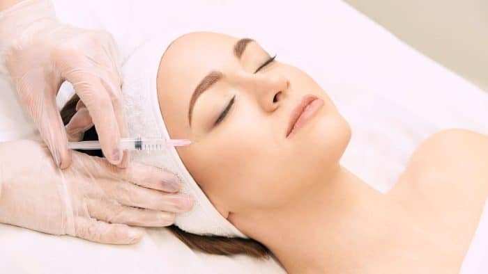 A person is lying down with their eyes closed, wearing a white headband. Gloved hands are seen administering an injection to their face, near the cheek area, suggesting a cosmetic or medical procedure. The setting appears clinical and professional.
