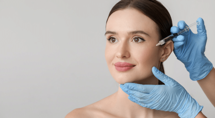 A woman with a bare shoulder is sitting and smiling slightly as a person wearing blue medical gloves gently holds her chin and cheek while preparing to give her a facial injection with a syringe. The background is plain and neutral.