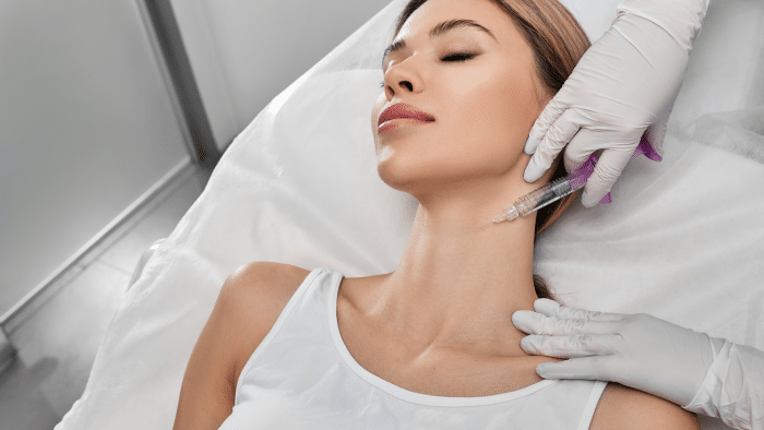 A woman lying on a medical bed with her eyes closed is receiving a cosmetic injection in her neck by a healthcare professional wearing white gloves. She appears calm, and the setting is clinical.