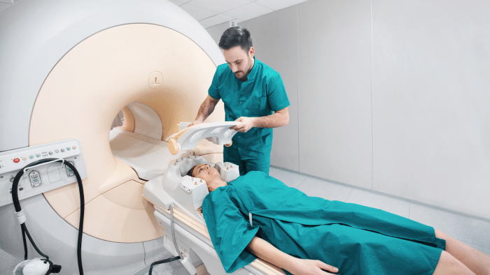 A medical professional in teal scrubs prepares a patient for an MRI scan. The patient, wearing a matching gown, lies on a table that will slide into a large, beige MRI machine. The room is clean and has bright, even lighting.