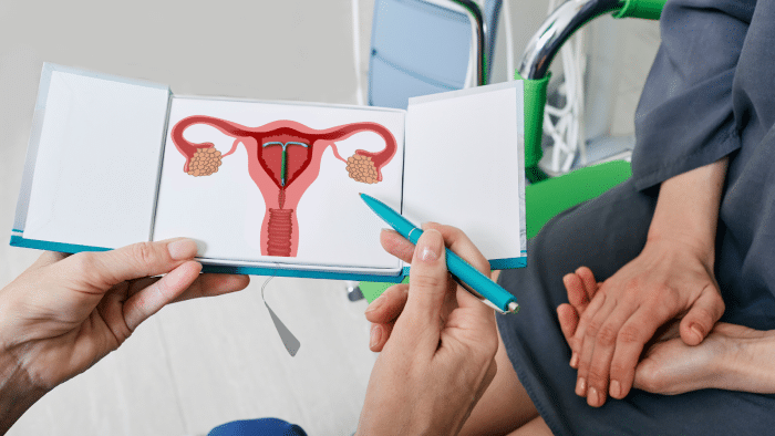 A person in a medical setting holds an anatomical illustration of the female reproductive system, pointing at it with a pen. Another person sits beside them with hands resting on their lap, listening attentively.