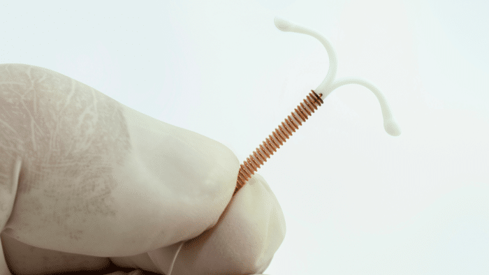 A gloved hand holds a small T-shaped copper intrauterine device (IUD) against a plain background. The IUD has a white plastic frame with copper wire coiled around its vertical stem and flexible arms at the top.