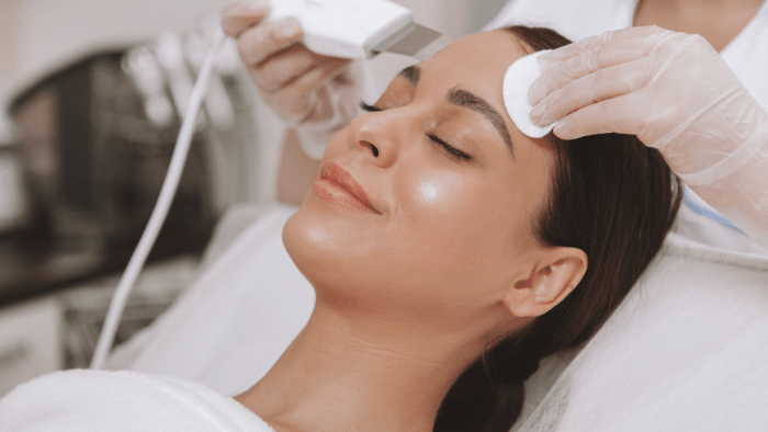 A woman lies with her eyes closed, receiving a skincare treatment. A gloved person is using an ultrasonic device on her forehead while holding a cotton pad on her skin. The woman appears relaxed and is lying on a treatment bed covered in a white towel.