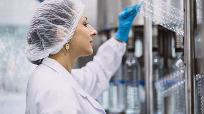 A person wearing a white lab coat, hairnet, and blue gloves inspects plastic bottles on a production line in a factory setting. Their profile is visible as they reach up to check the bottles, highlighting their focused and attentive expression.