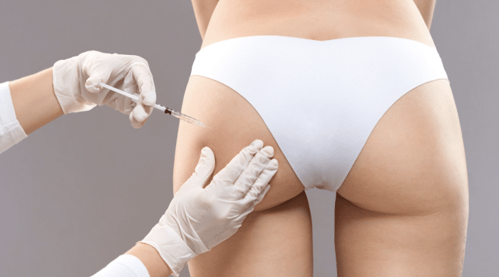 A person wearing white undergarments is receiving an injection in the buttock from a professional wearing white gloves. The background is neutral gray.