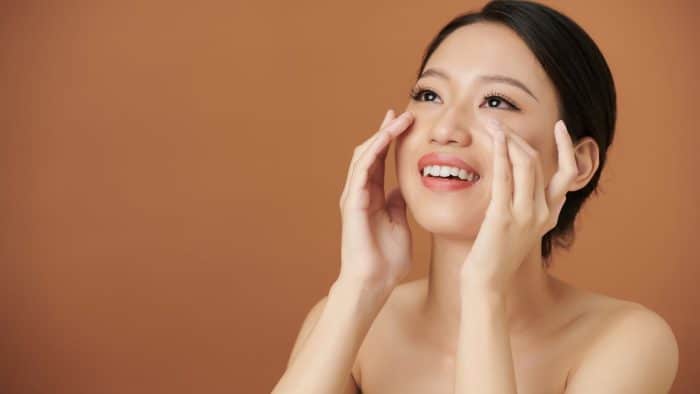 A woman with dark hair smiles, gently touching her face with both hands. She has a radiant complexion and is wearing minimal or no makeup. The background is a solid brown color.