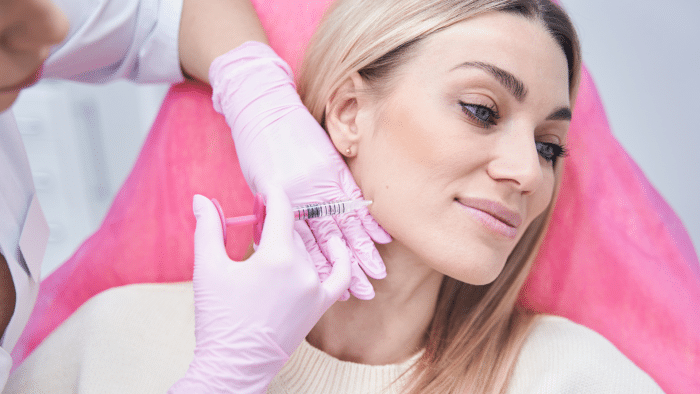 A woman with blonde hair receives a cosmetic injection on her jawline from a medical professional. She is wearing a white top, and the medical professional is wearing pink gloves. The woman appears relaxed in a clinical setting.