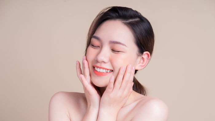 A woman with smooth skin and dark hair smiles with her eyes closed, gently touching her face with both hands. She appears content and relaxed, with a neutral beige background highlighting her serene expression.