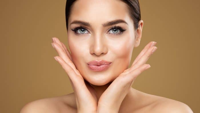 A person with fair skin and dark hair is posing against a light brown background. Their hands are placed under their cheeks, framing their face. They have striking blue eyes, neatly groomed eyebrows, eyeliner, and nude lipstick, with a calm expression.