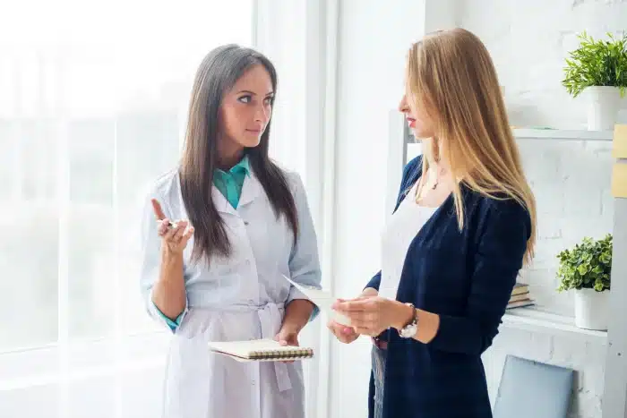 A female doctor in a white coat holding a clipboard is talking to a woman with long blonde hair wearing a dark cardigan. They are standing by a window with shelves and potted plants in the background.