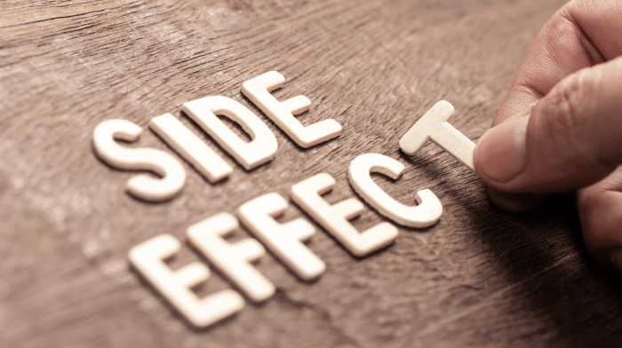 A hand is placing the last letter "T" to complete the phrase "SIDE EFFECT" formed with wooden letters on a wooden surface.