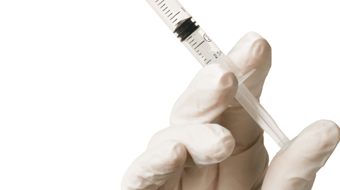 A gloved hand holds a syringe filled with liquid against a plain white background. The syringe is positioned with the needle pointing upwards and the measurement markings are visible on the side.