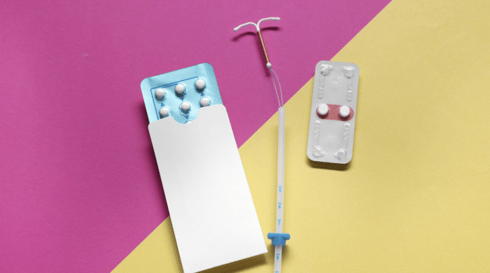 A photo showing various forms of contraception on a pink and yellow background. There is a blister pack of white pills partially inside a white envelope, an intrauterine device (IUD) with an insertion tool, and a smaller blister pack with two pills.