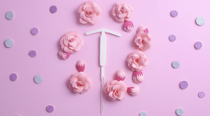 A white IUD is placed in the center of a pink background decorated with pink paper flowers and purple glitter circles, forming a visually appealing and decorative arrangement.